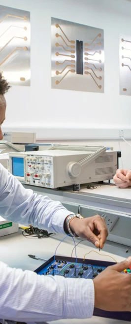A lecturer and a student in an electronics lab working on a circuit board experiment. The lecturer is smiling while overseeing the student’s work. A lecturer and a student in an electronics lab working on a circuit board experiment. The lecturer is smiling while overseeing the student’s work.